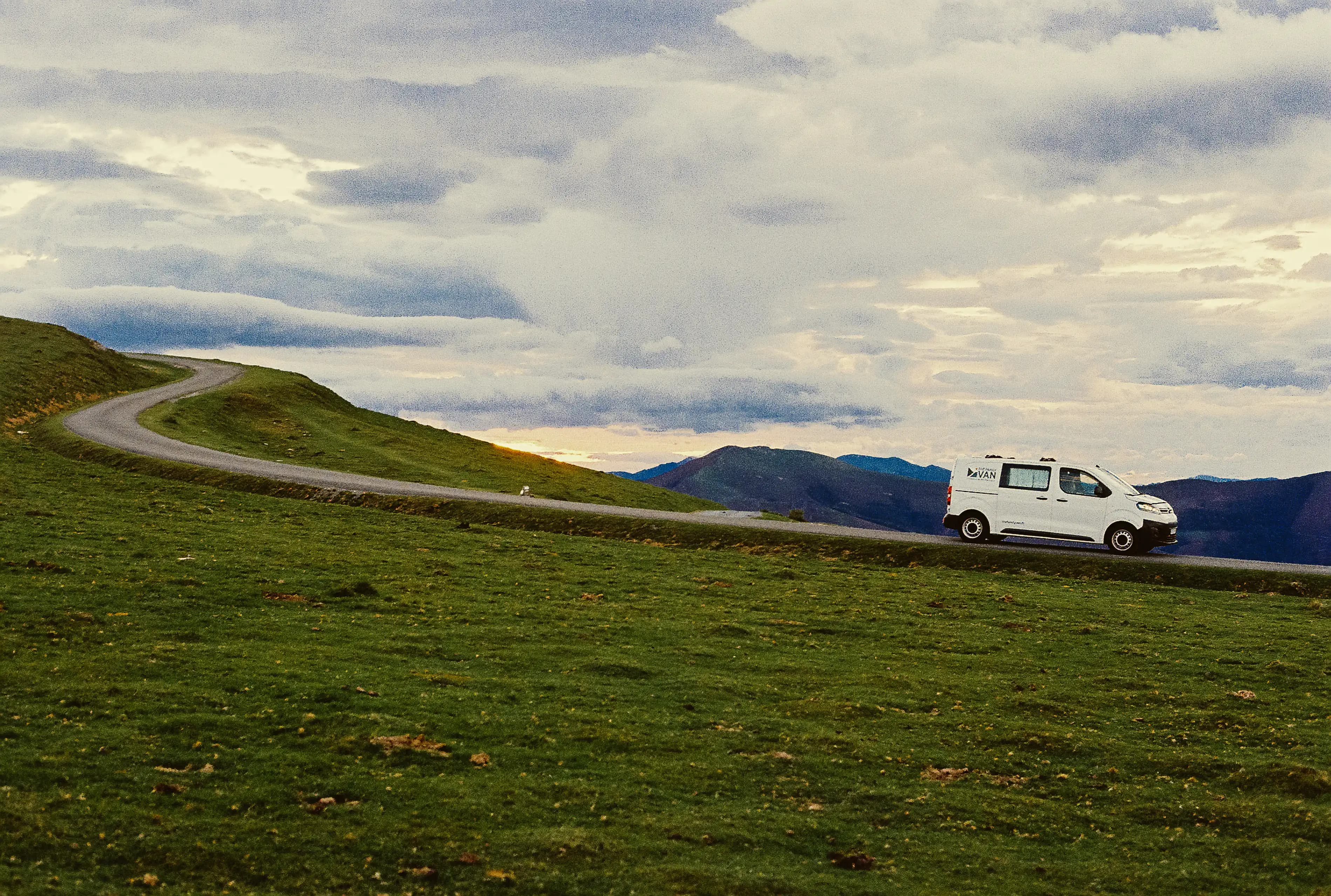 Campervan blanc sur une route de crête herbeuse, nuages bas au-dessus des montagnes