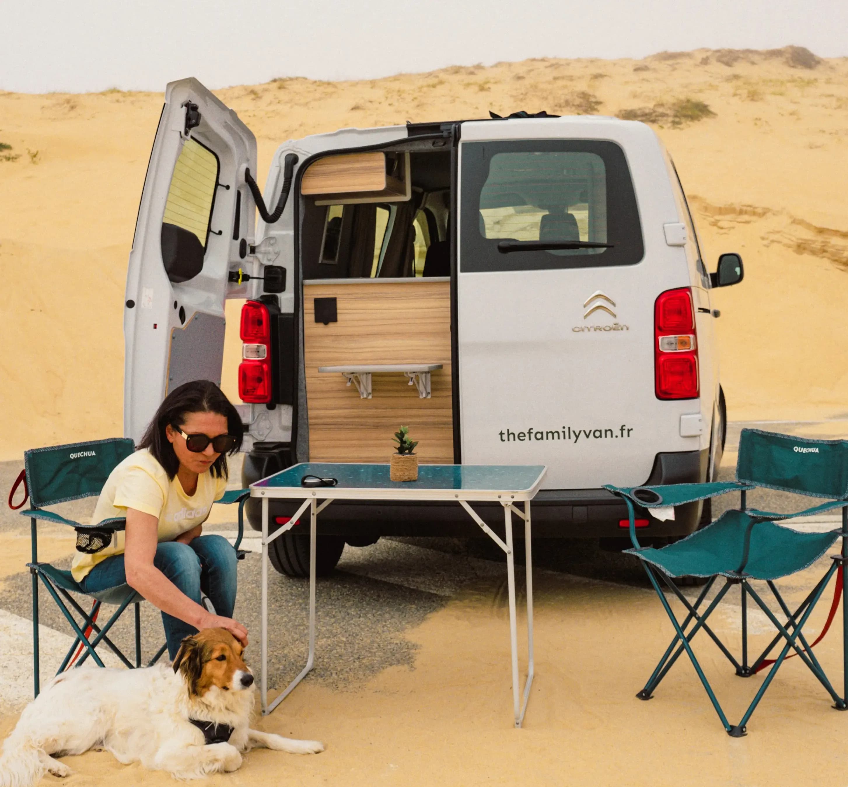 Une femme caresse son chien près d'un campervan portes arrière ouvertes, au milieu des dunes