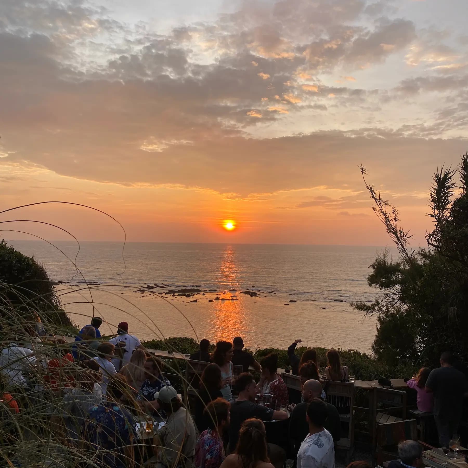 Foule attablée à un bar en plein air sur la falaise devant un coucher de soleil sur l'océan