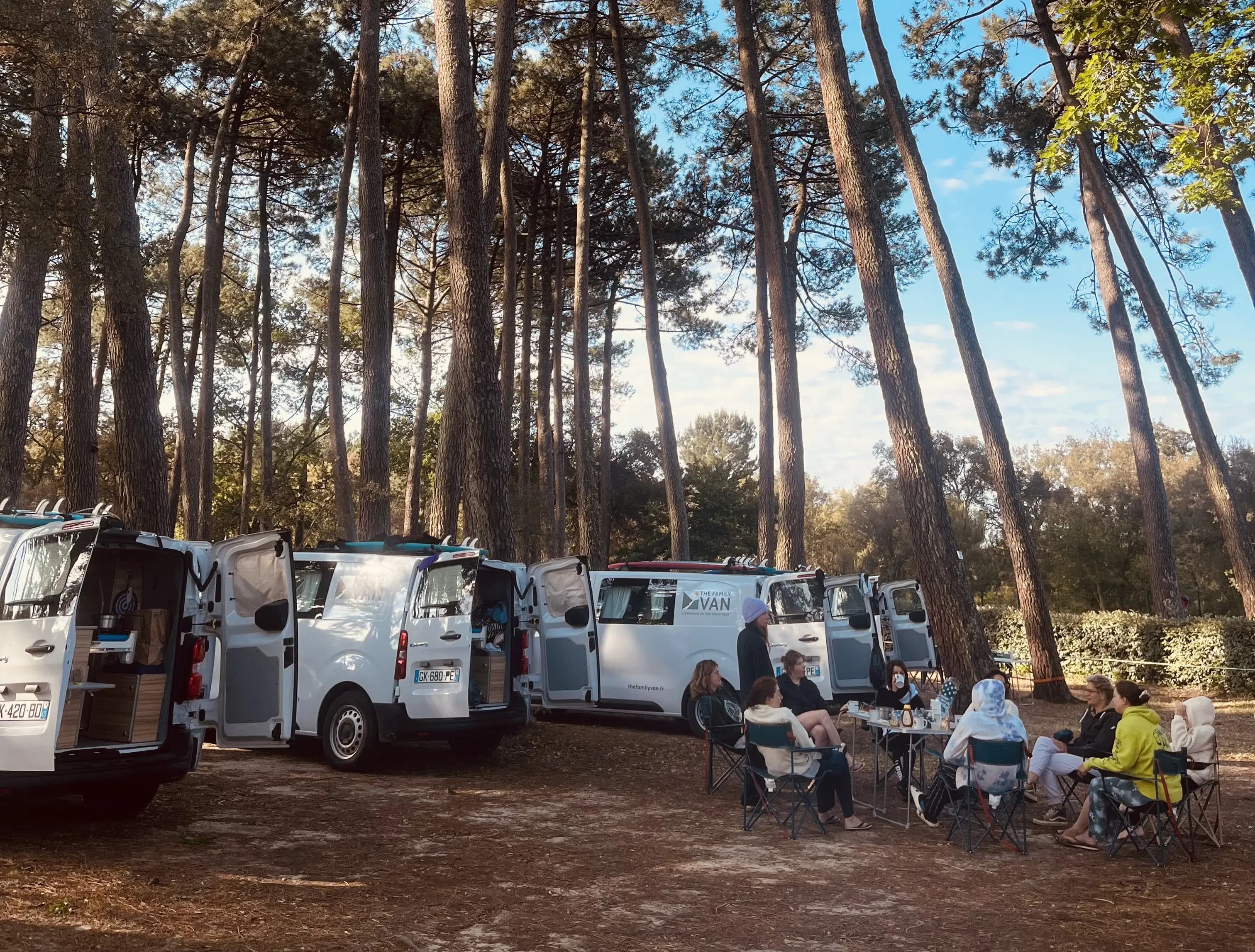Plusieurs campervans alignés sous les pins, groupe attablé en chaises de camping devant les vans