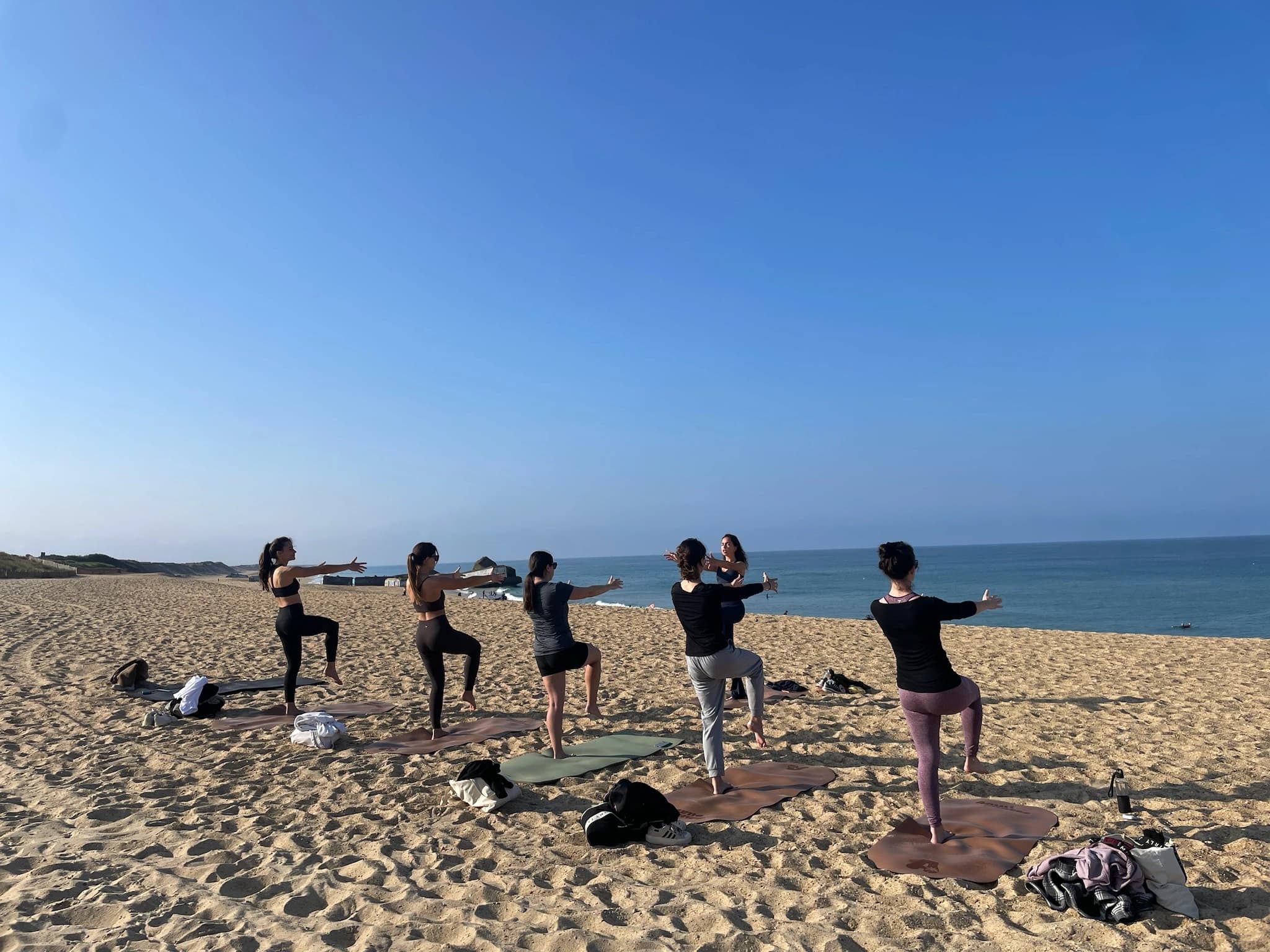 Cinq participantes en équilibre bras écartés sur des tapis face à l'océan, plage déserte