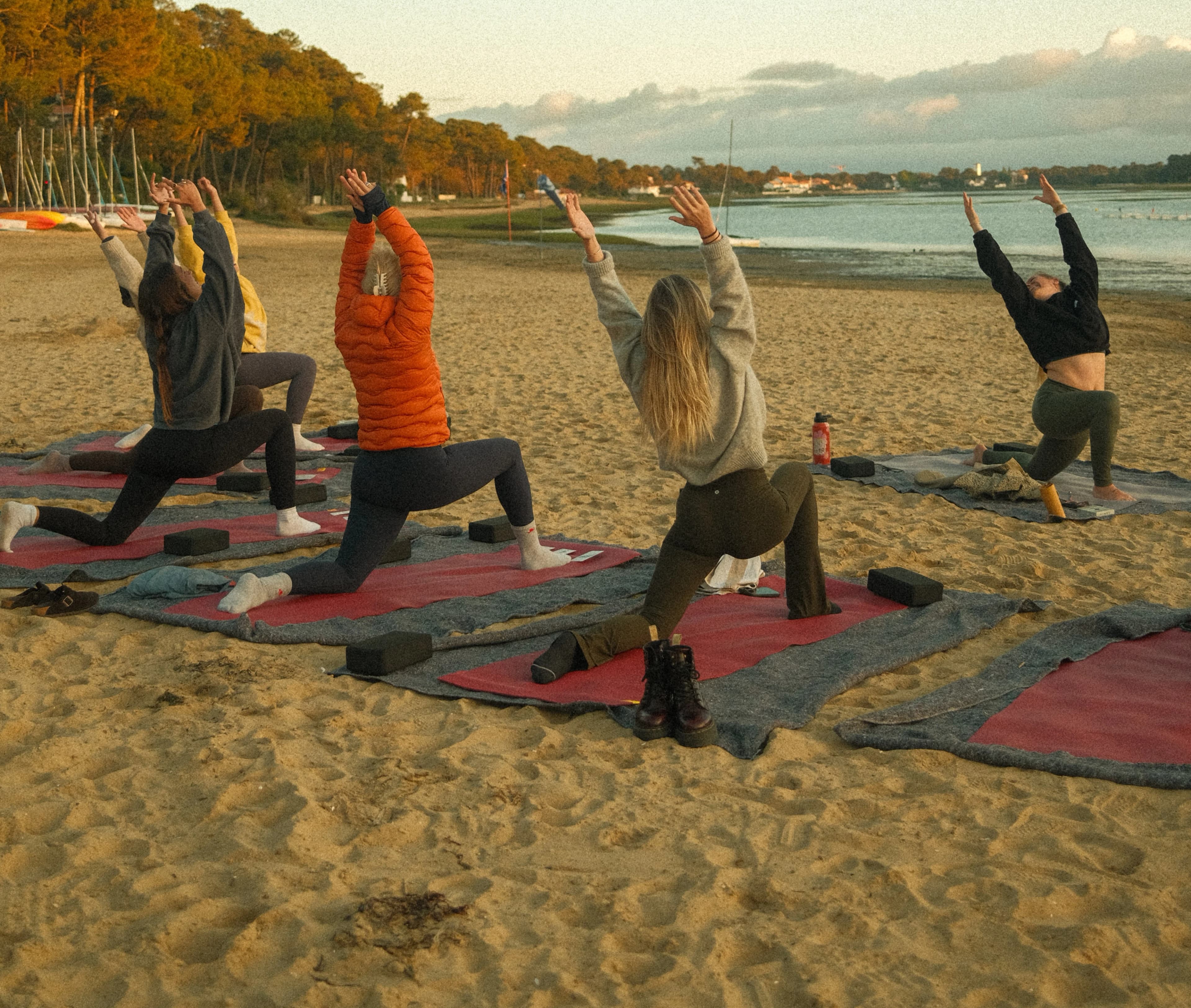 Cours collectif de yoga sur le sable au couchant, fente basse bras levés vers le ciel