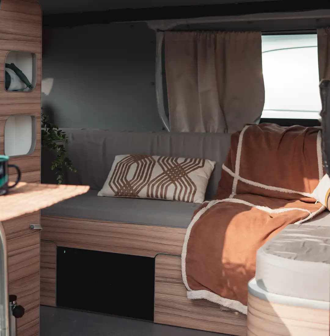 Banquette du van habillée d'un plaid terracotta et de coussins, rideaux et bois clair