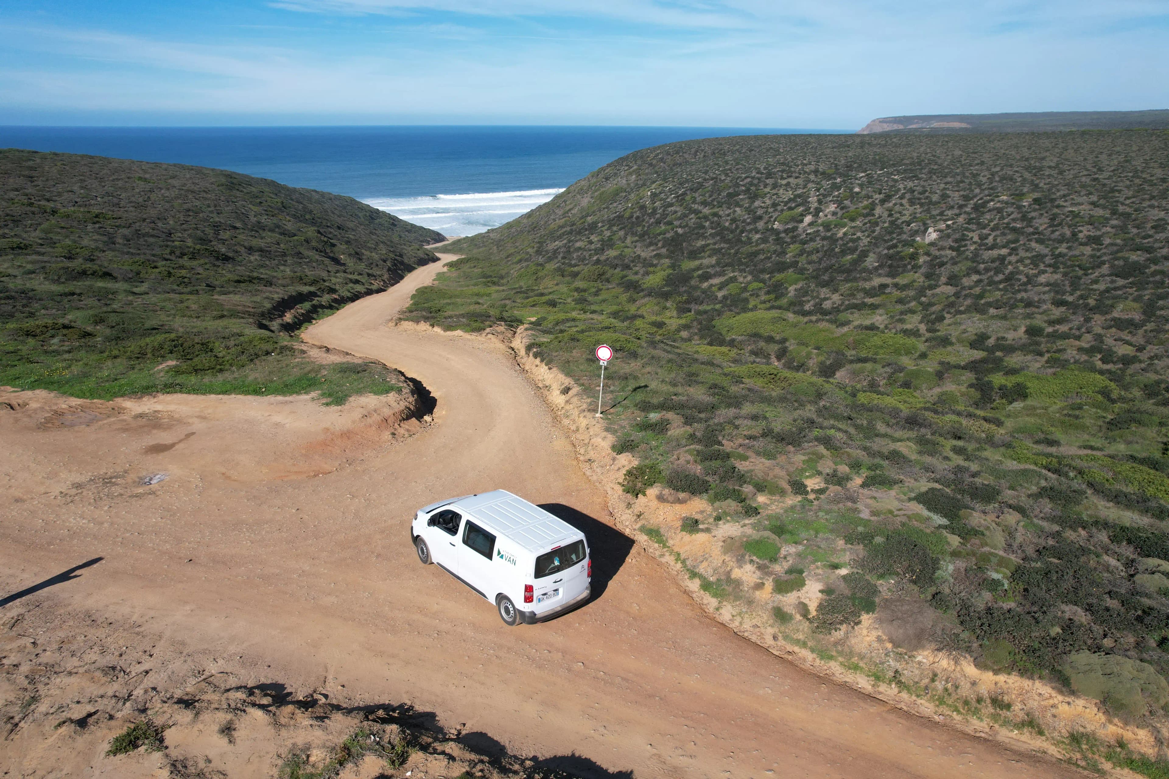 Van blanc sur une piste de terre serpentant entre les collines jusqu'à une plage de surf