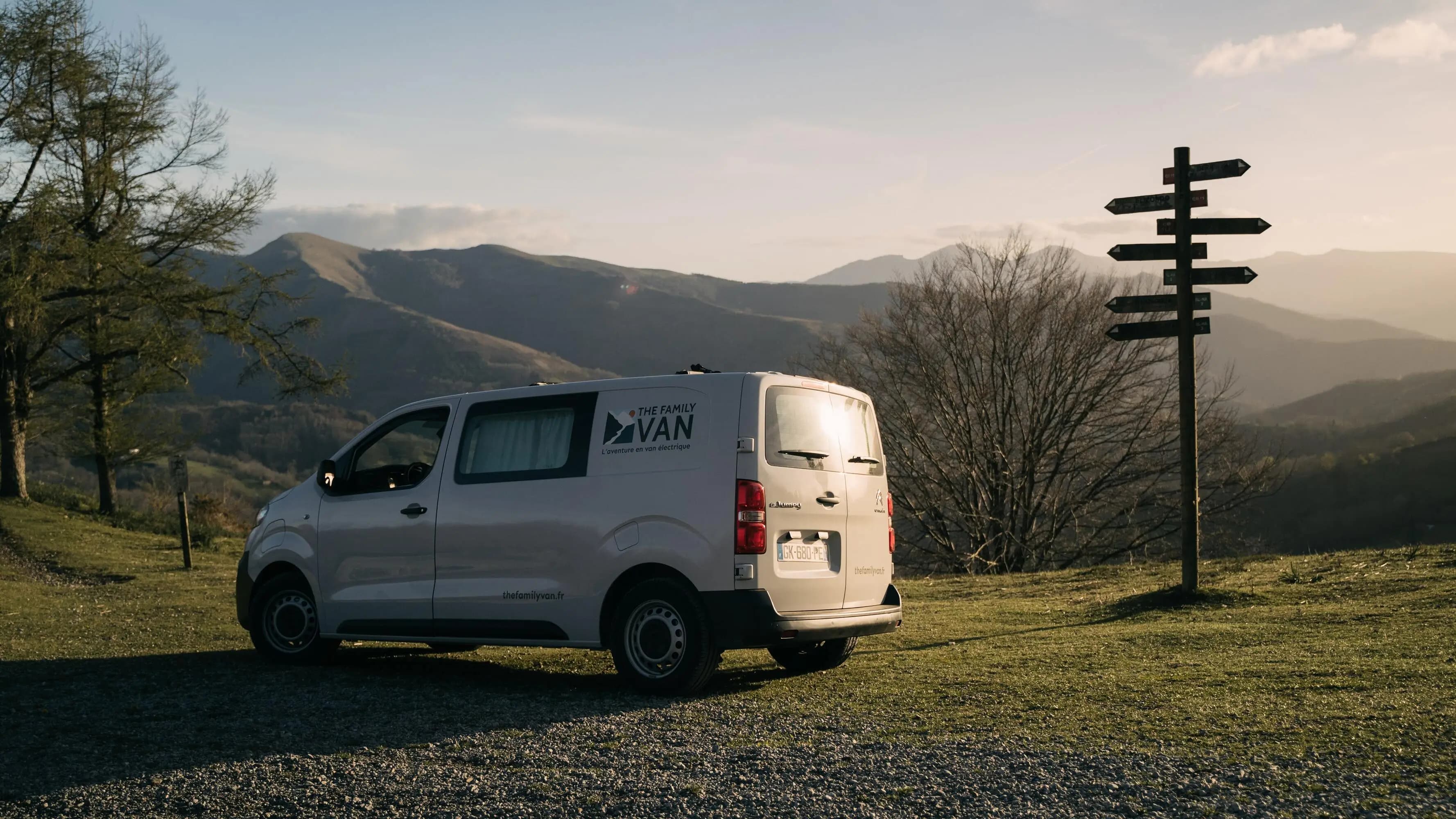 Campervan blanc garé sur un col herbeux près d'un poteau directionnel, montagnes au couchant