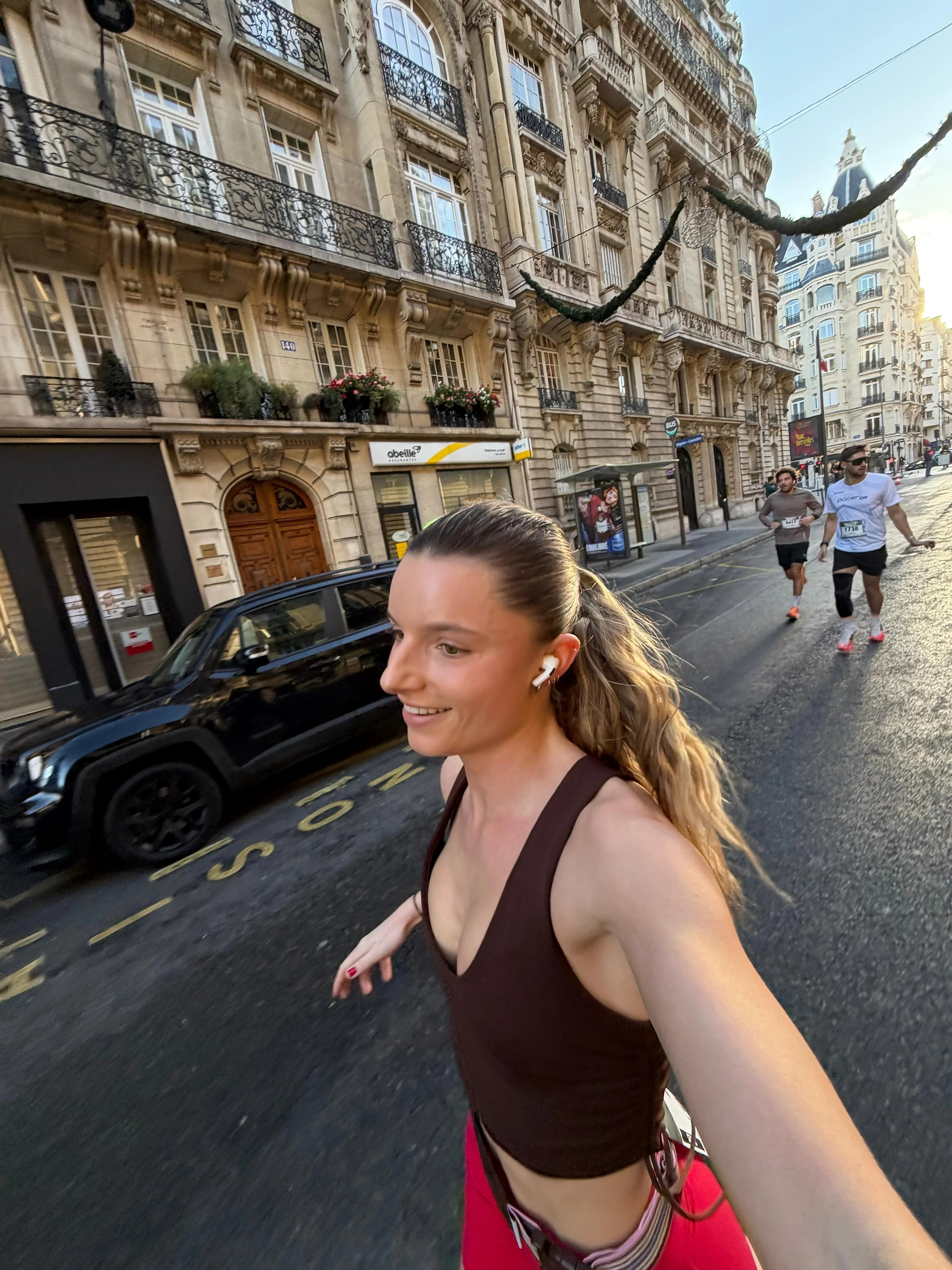 Coureuse souriante en selfie dans une rue haussmannienne, deux coureurs derrière elle