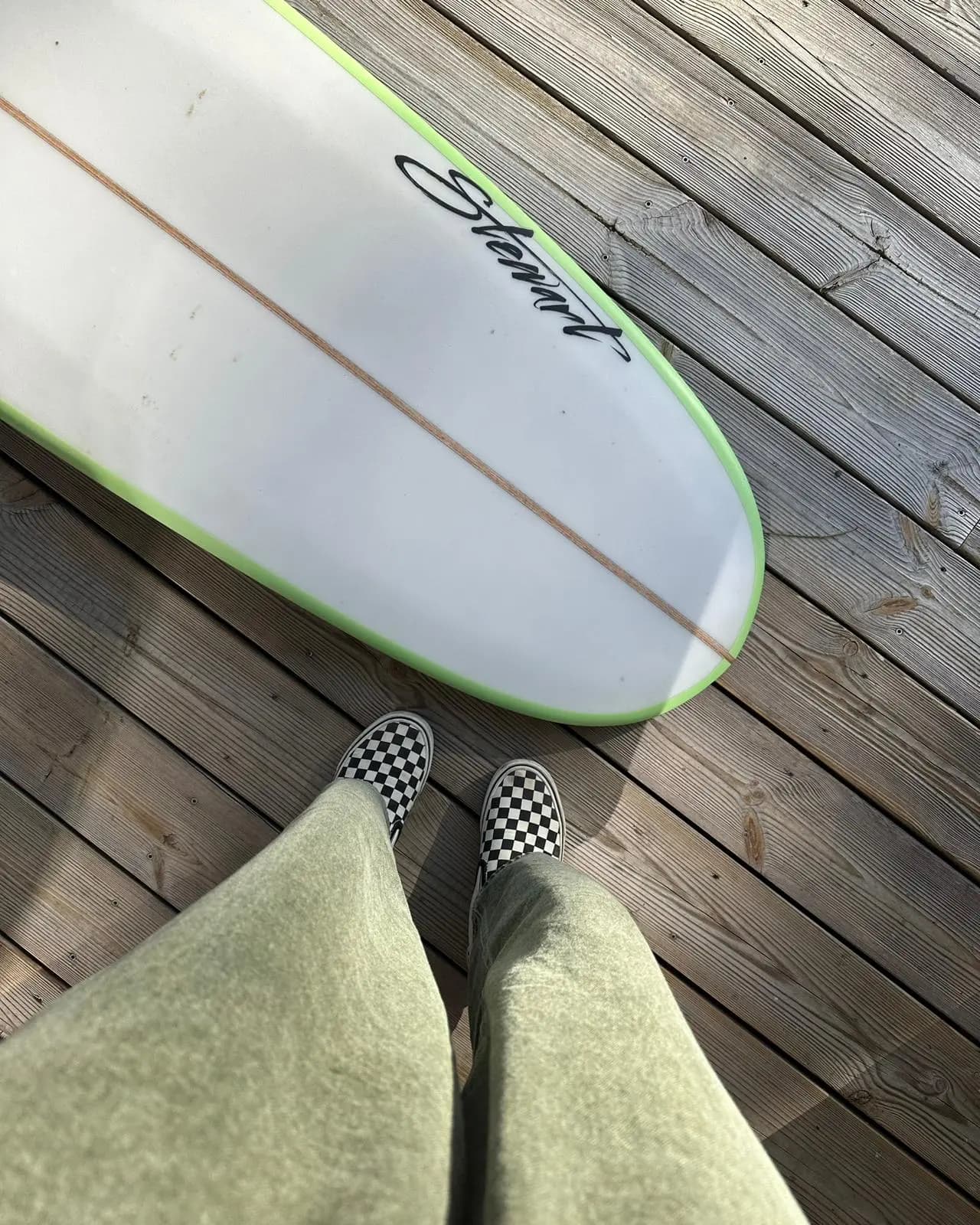 Planche de surf posée sur une terrasse en bois, jambes et baskets à damier au premier plan