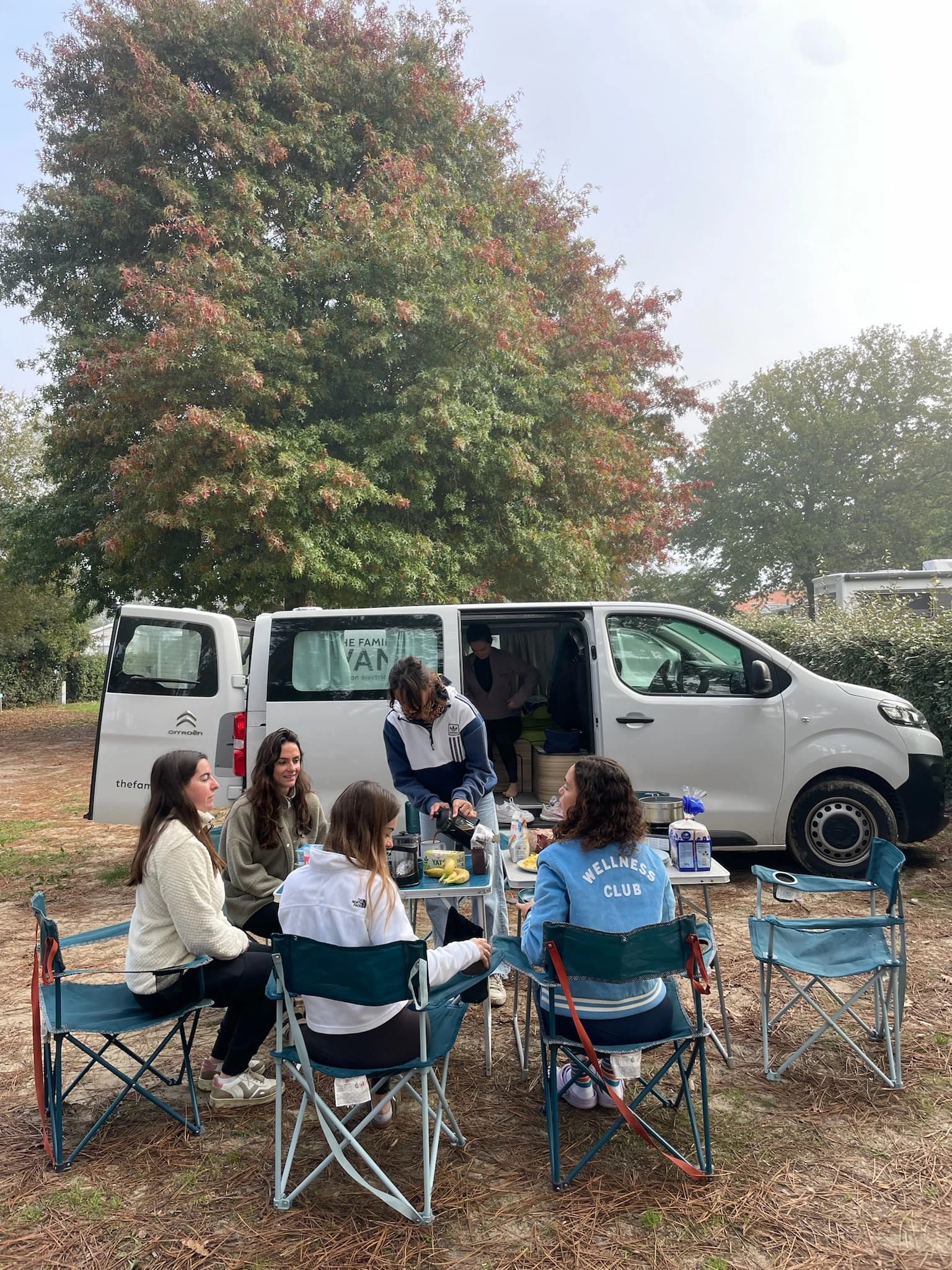 Petit-déjeuner entre femmes à une table de camping devant un van blanc, arbre d'automne derrière