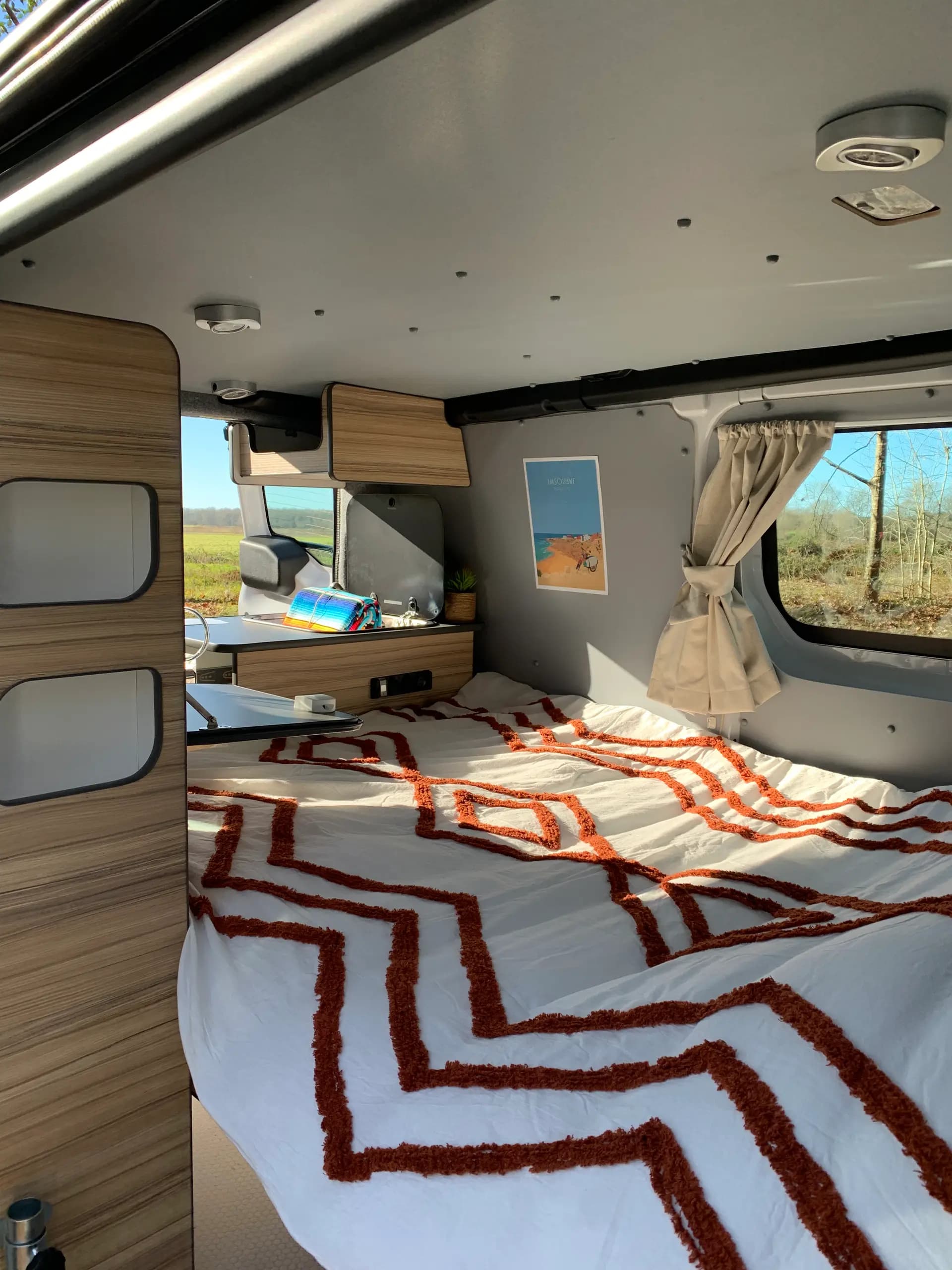 Grand lit fait dans un campervan, couvre-lit blanc à motifs orange et rideaux tirés à la fenêtre