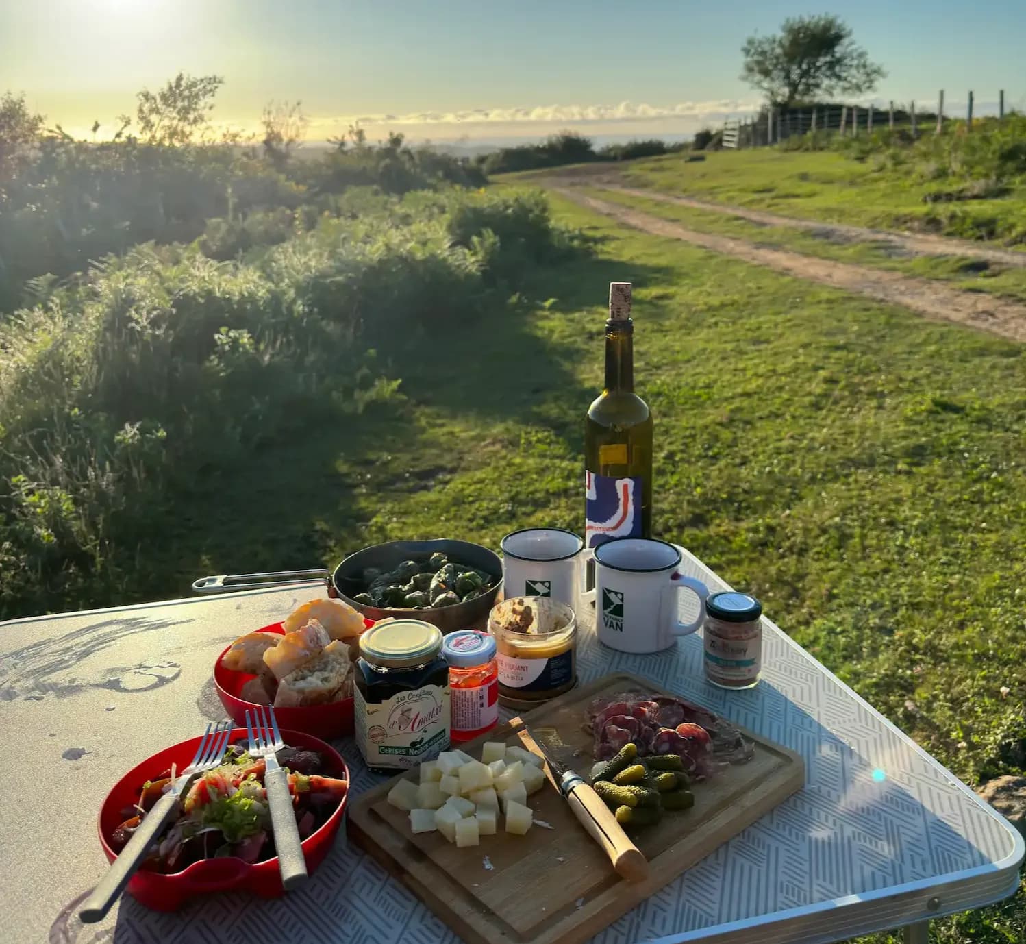 Table de camping garnie de fromage, charcuterie et vin blanc face à un chemin de campagne