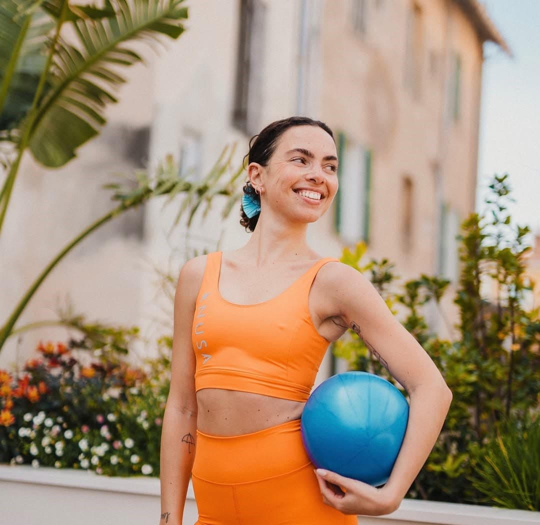 Coach de pilates en tenue orange, un ballon bleu sous le bras, souriante dans un patio fleuri